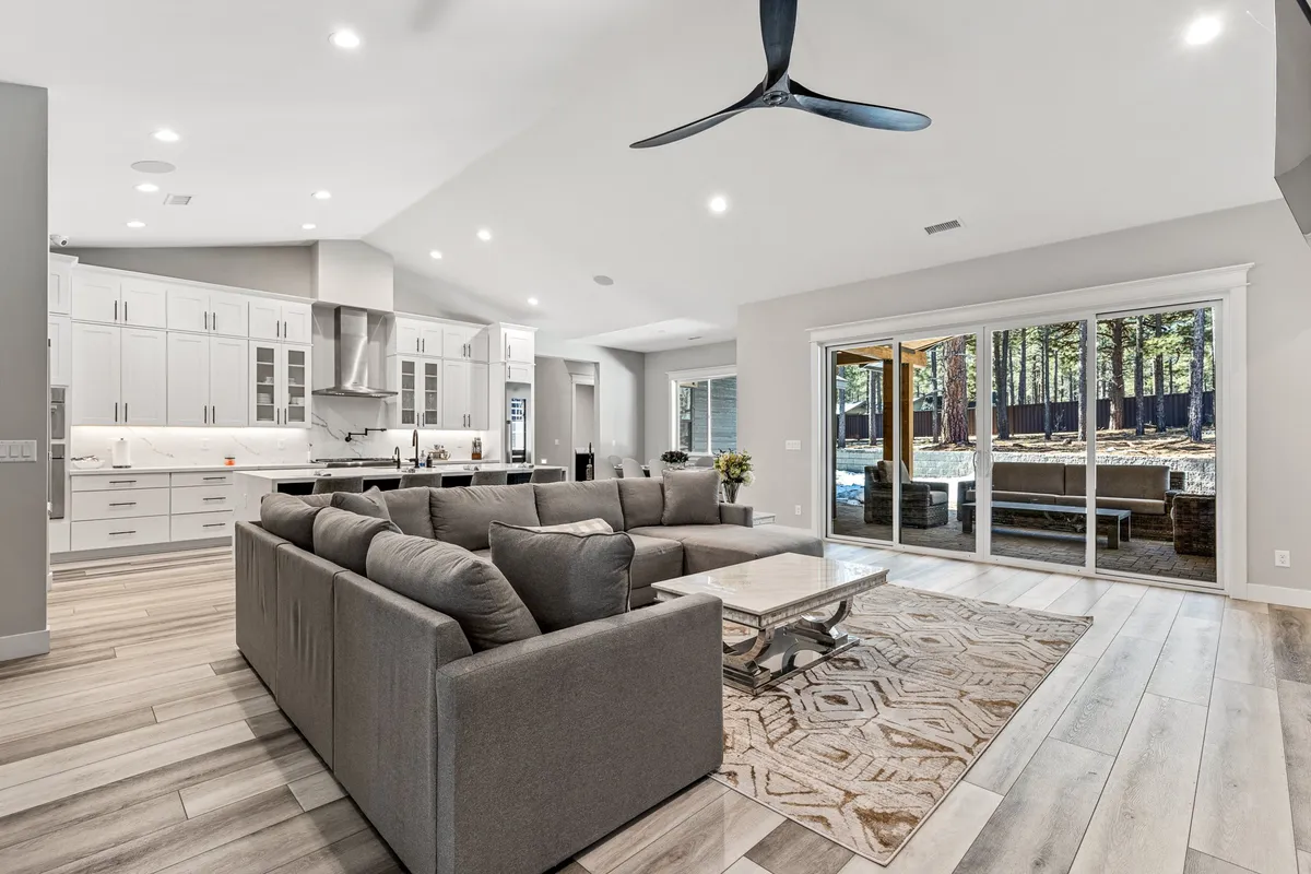 This image captures a spacious open-plan living area featuring a large sectional sofa, a modern kitchen, and floor-to-ceiling windows that flood the space with natural light. The inviting living room flows seamlessly into the kitchen, and the expansive windows reveal a scenic outdoor view, enhancing the sense of mountain serenity and openness.