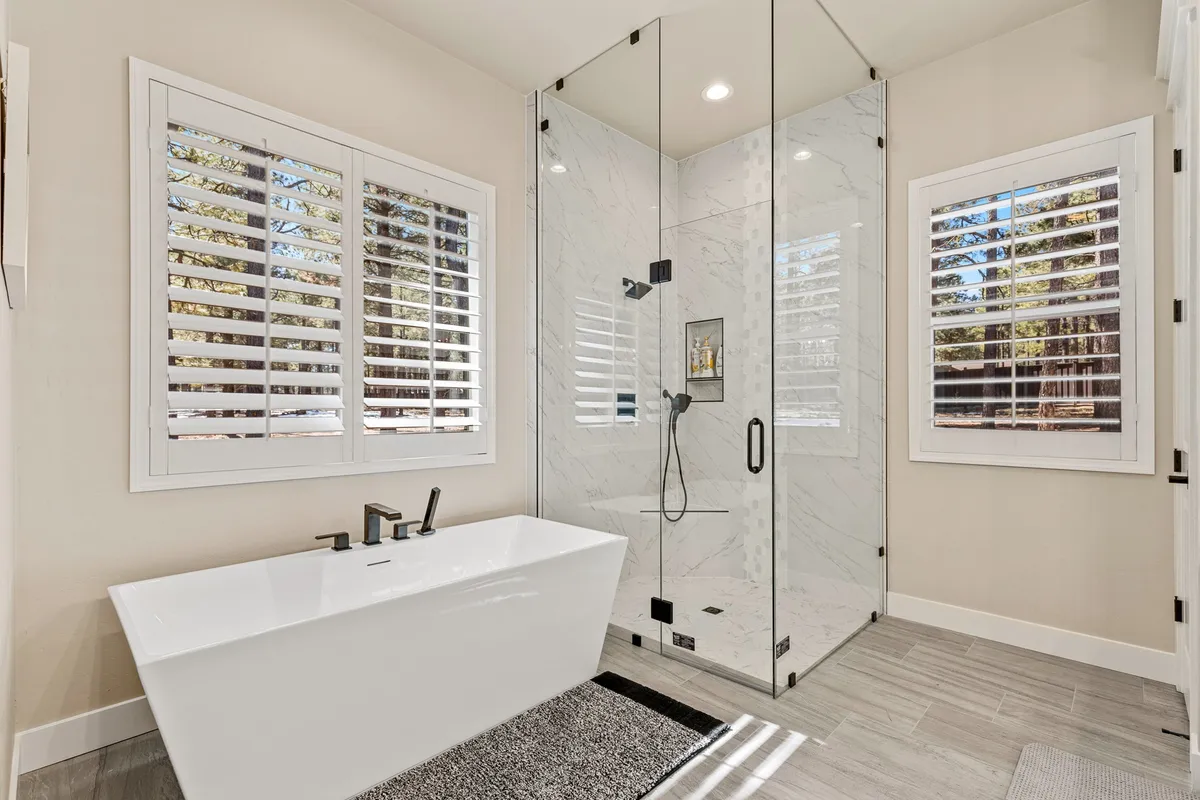 A luxurious bathroom featuring a freestanding bathtub, modern glass shower enclosure, and a window with shutters for privacy. Natural light enhances the spa-like atmosphere.