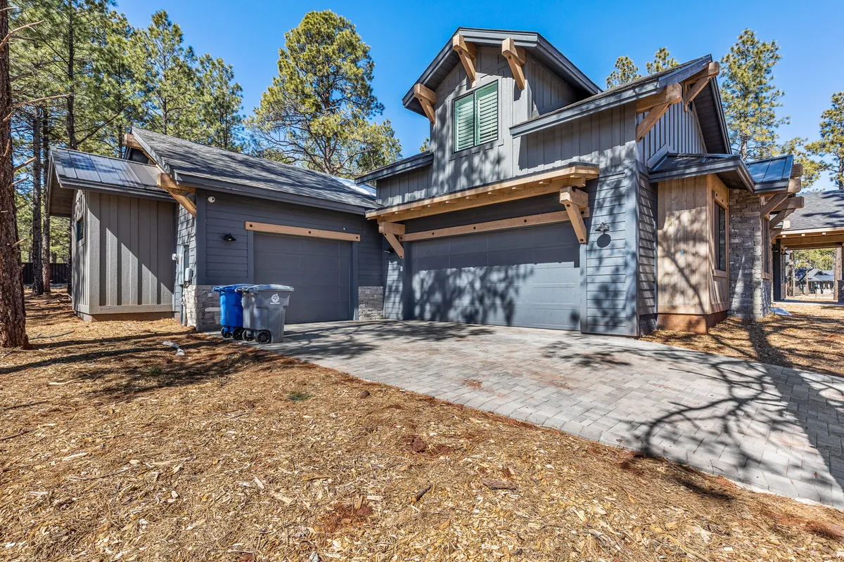 A view of the exterior with garage doors, stone accents, wooden beams, and tall pine trees framing the property. The setting suggests ample outdoor space for family gatherings and playtime, with natural elements blending into the landscape.