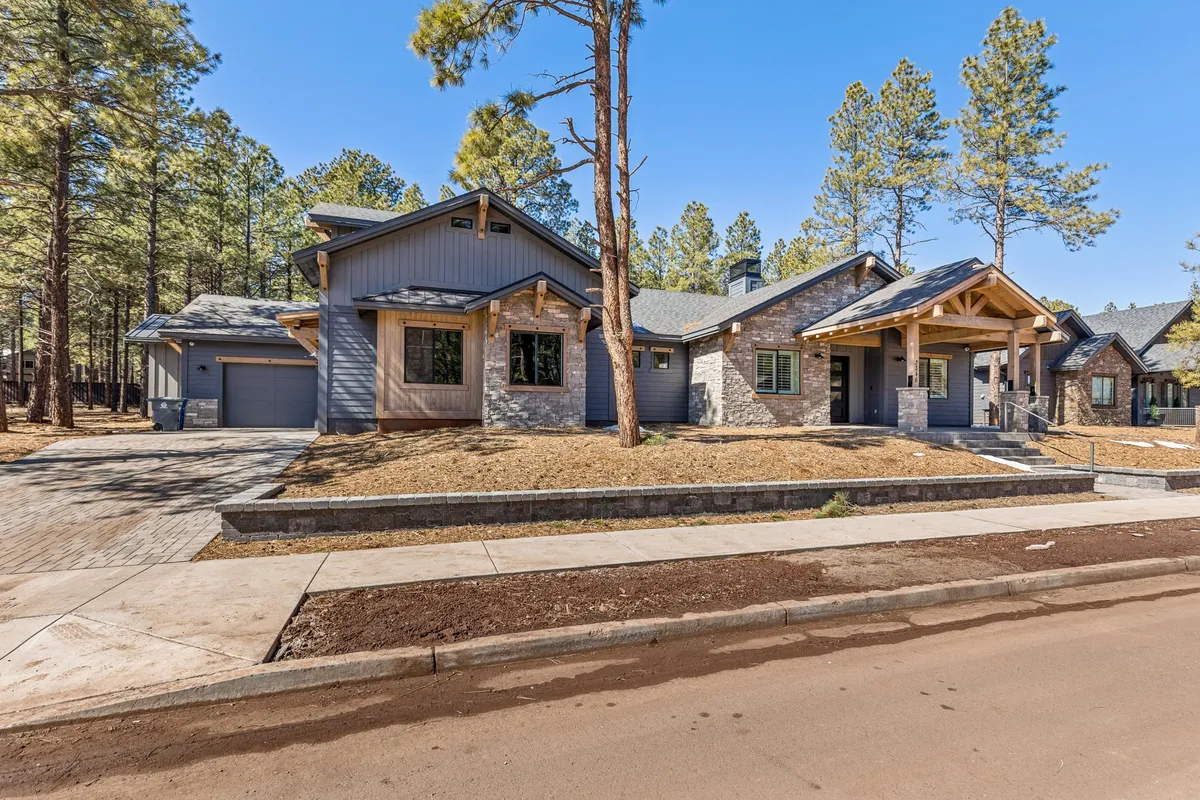 The image shows a welcoming exterior view of the home's front porch, stone facade, and large windows, all framed by tall pine trees. The scene glows with sunset light, suggesting both relaxation and the beauty of Flagstaff's natural surroundings.