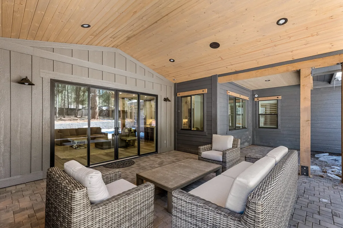 An inviting patio with woven seating, a wooden ceiling, and large glass doors that seamlessly connect inside and outside living. The backyard and surrounding nature are visible, highlighting the blend of comfort and forest environment.