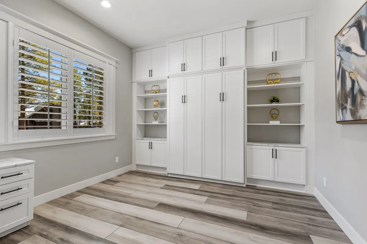 A peaceful, minimalist space featuring a large white cabinetry, a window with plantation shutters letting in natural light, wooden floor tiles, and decorative shelving. The setup includes a writing desk ideal for working, reading, or studying.